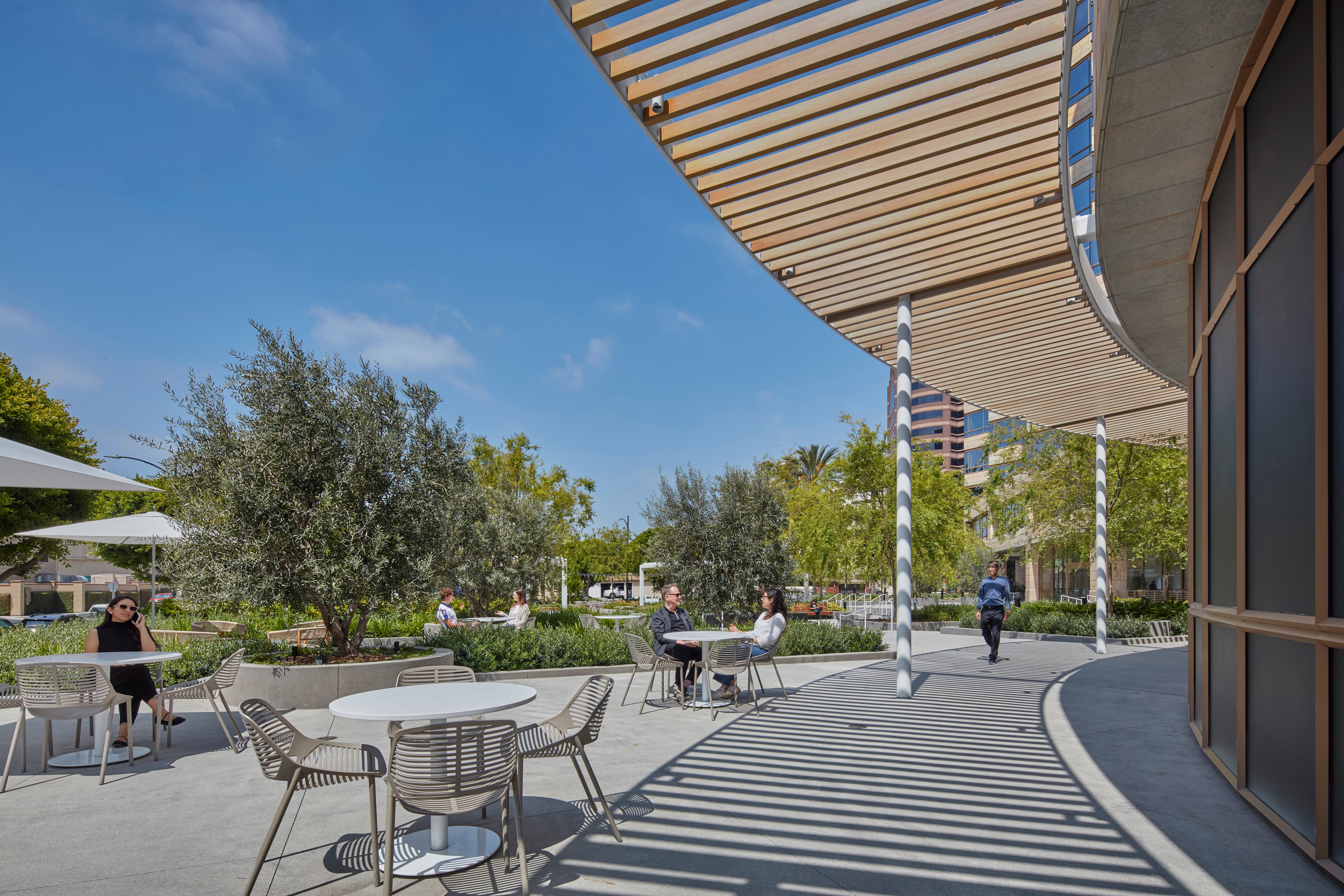 Studio Plaza outdoor seating area under a wood pergola, with people relaxing and walking by.