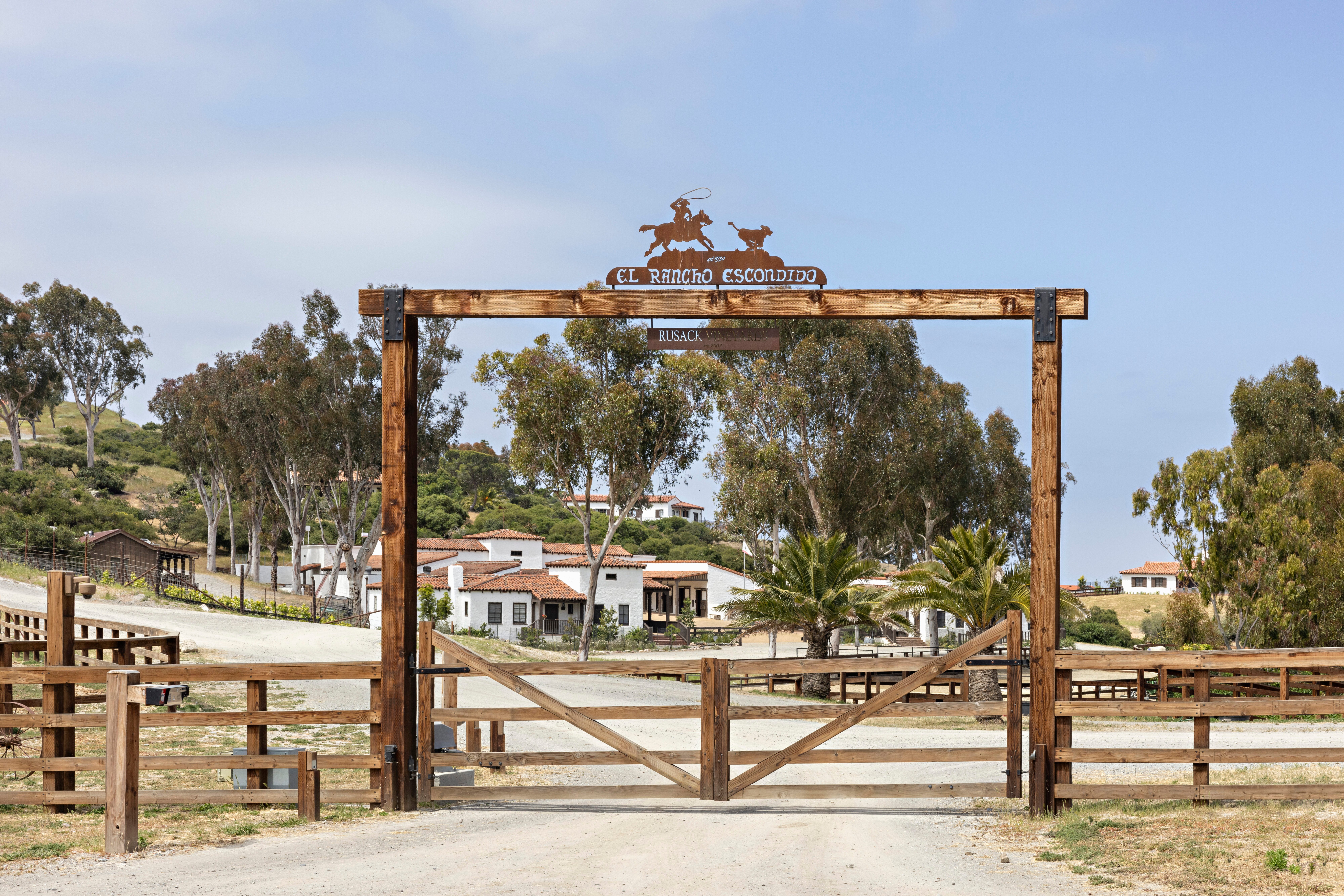 Wooden entrance gate with 'El Rancho Escondido' sign, leading to ranch buildings.