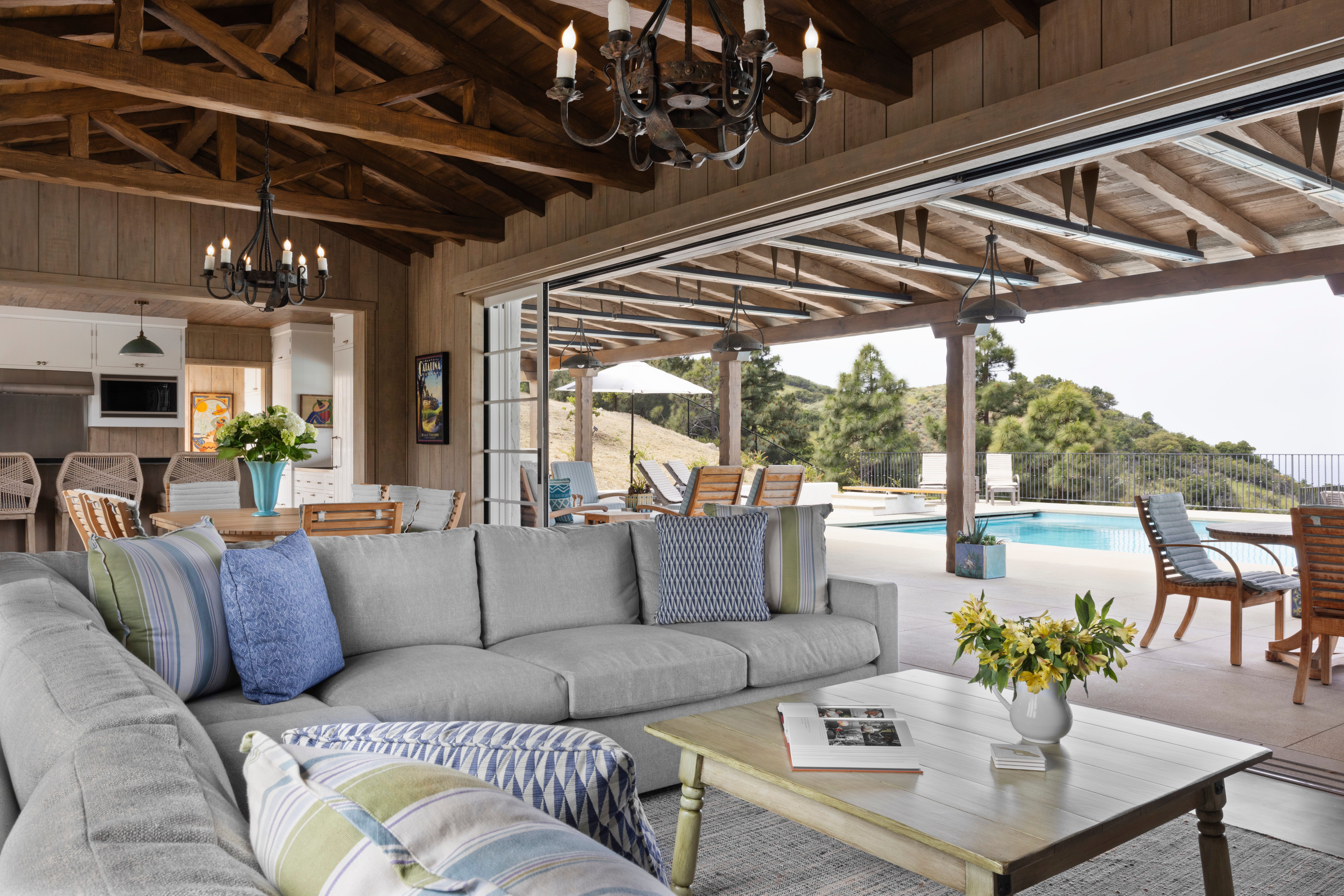 Living room with wood-beamed ceiling and gray sectional opens to a pool and hillside view at El Rancho Escondido, Catalina Island.